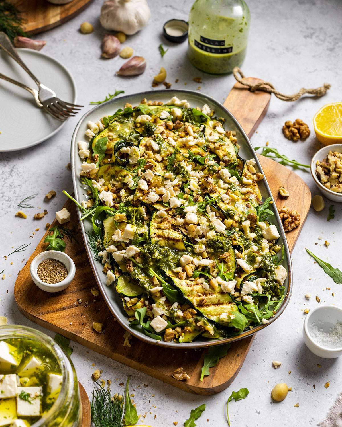 Vegan Grilled Zucchini Salad with walnuts and vegan feta crumbles on a serving platter, sitting on a white dinner table surrounded by ingredients.