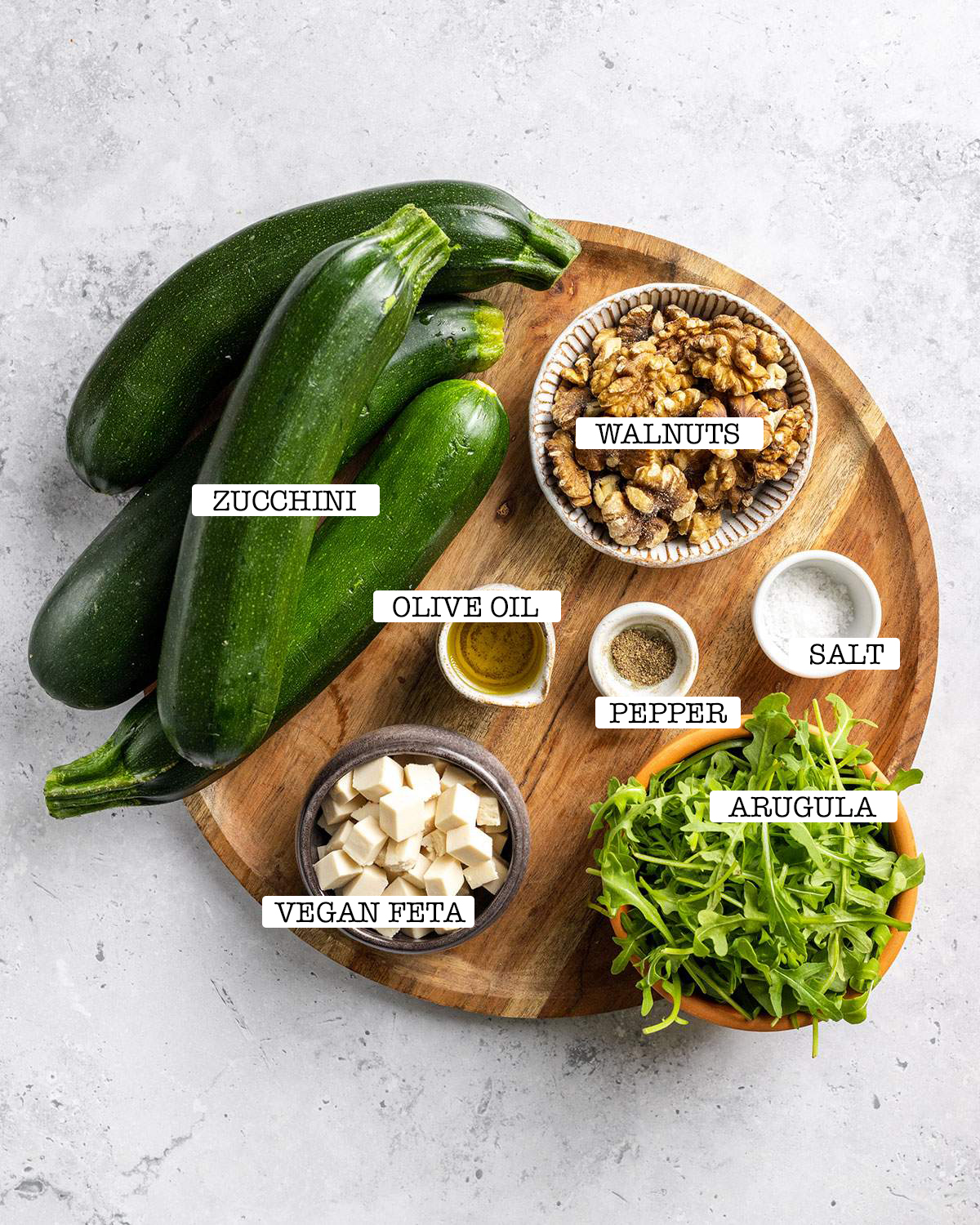 The ingredients for grilled zucchini salad with walnuts and vegan feta are sitting on a wooden tray with labels.