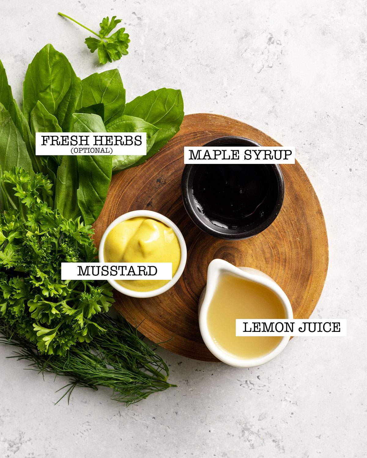 The ingredients for a simple oil free dressing are sitting on a white table. Small measured jugs with lemon juice, Dijon mustard and maple syrup are sitting on a round wooden board and a handful of fresh herbs are arranged besides it.