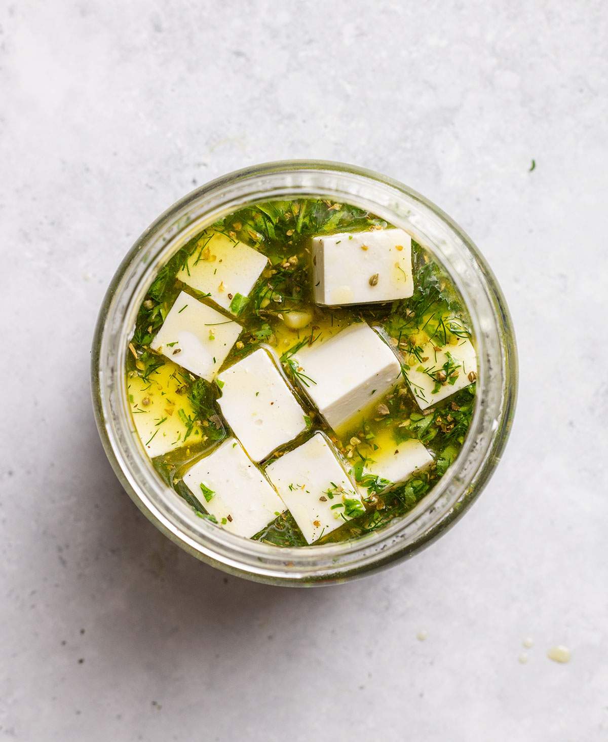 Vegan feta cubes are sitting in a oily and herby marinade in a glass jar.
