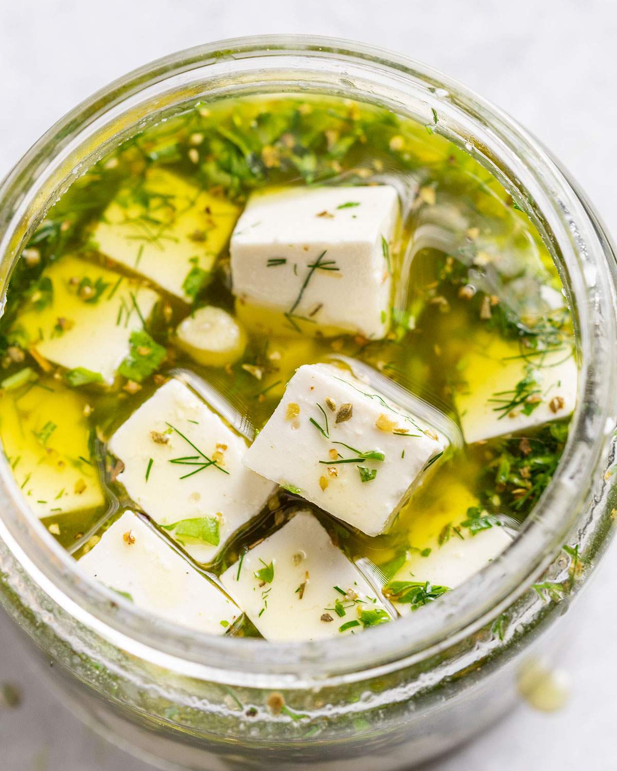 Vegan feta cubes are sitting in a oily and herby marinade in a glass jar.