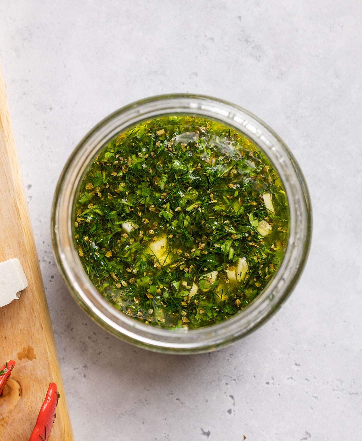 Vegan Feta marinade can be seen in a glass jar with lots of finely chopped herbs inside the marinade.