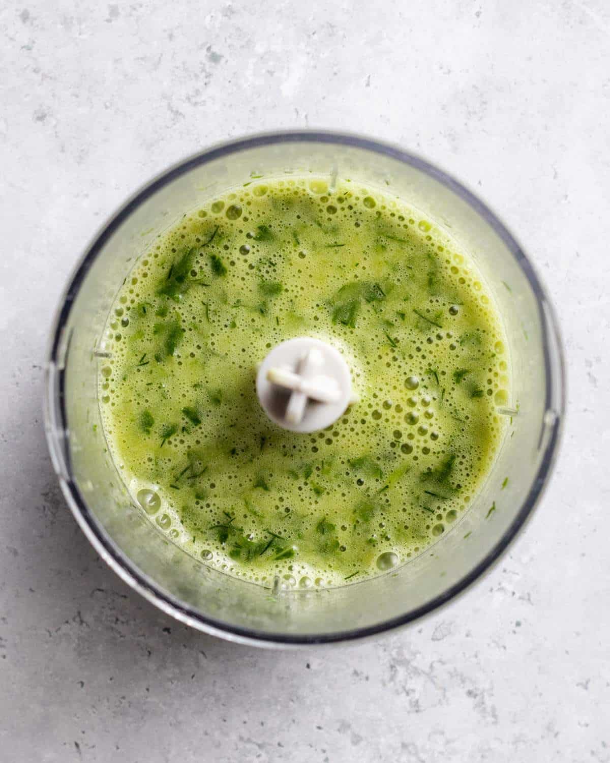 A small blender bowl is shown from above with a green salad dressing inside. The herbs are finely chopped up in the dressing.