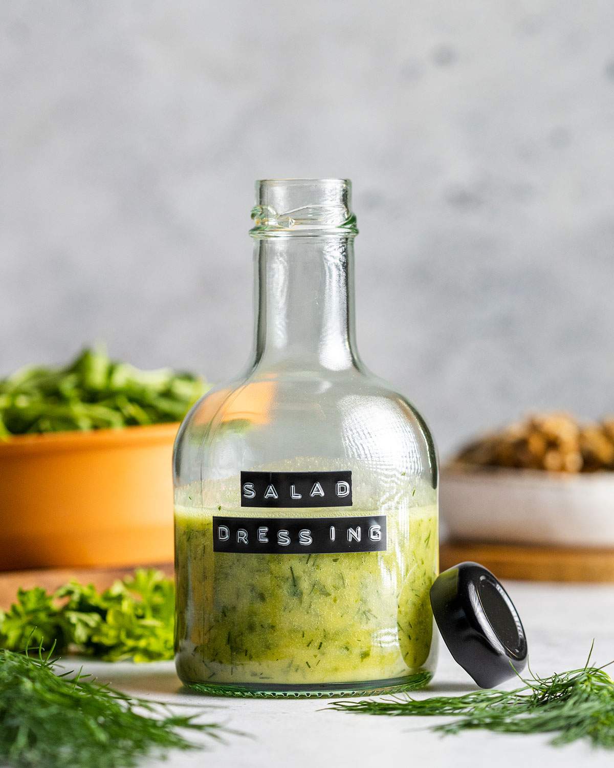 An open glass bottle with a narrow neck and a black label reading 'salad dressing' is sitting on a table surrounded by fresh herbs. The black lid for the bottle is leaning against the side.