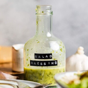 A small glass bottle with a label reading 'salad dressing' is sitting on a table in between dishes. There is a green dressing inside the bottle and you can see small pieces of chopped herbs.