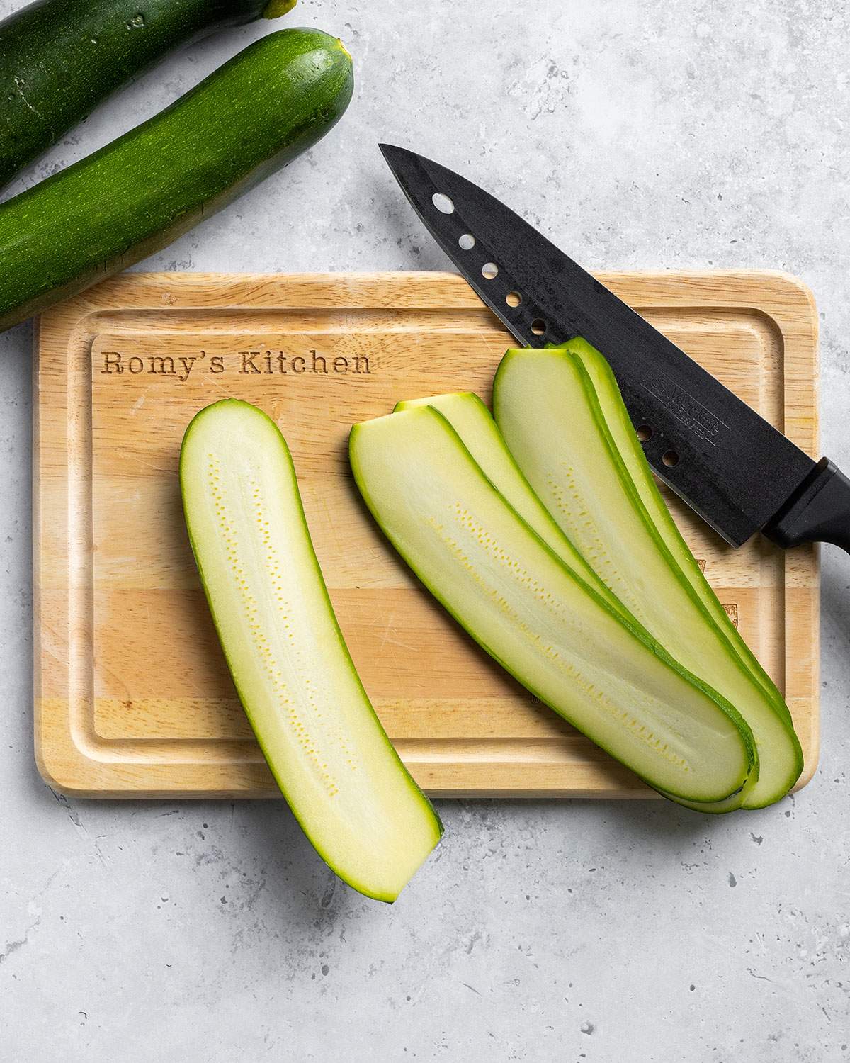 Courgette has been sliced and the slices are laying on a wooden board next to a large kitchen knife.