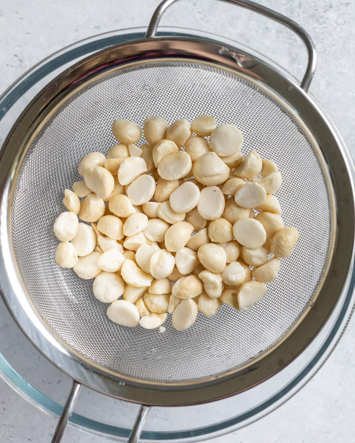 Soaked and rinsed macadamia nuts are seen in a colander above a glass bowl.