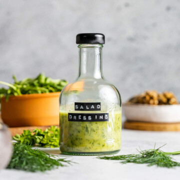 A bottle of oil free salad dressing is sitting on a white table with fresh herbs and other salad ingredients surrounding.