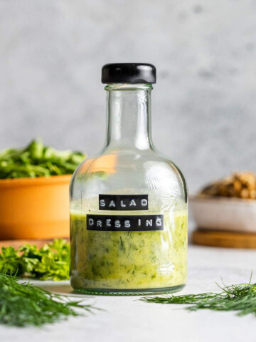 A bottle of oil free salad dressing is sitting on a white table with fresh herbs and other salad ingredients surrounding.