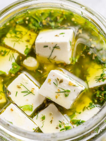 An open jar of vegan feta in a herby marinade is shown up close.