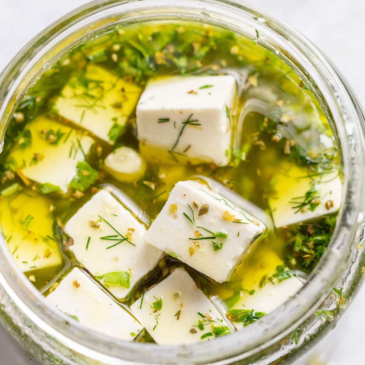 An open jar of vegan feta in a herby marinade is shown up close.