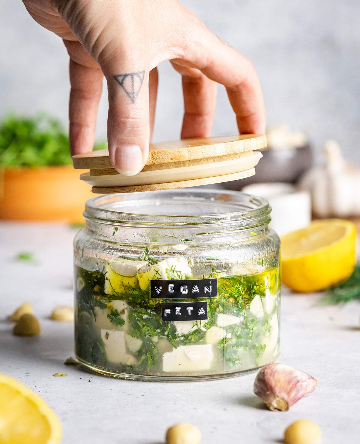 A hand is opening a glass jar filled with vegan feta and marinade.