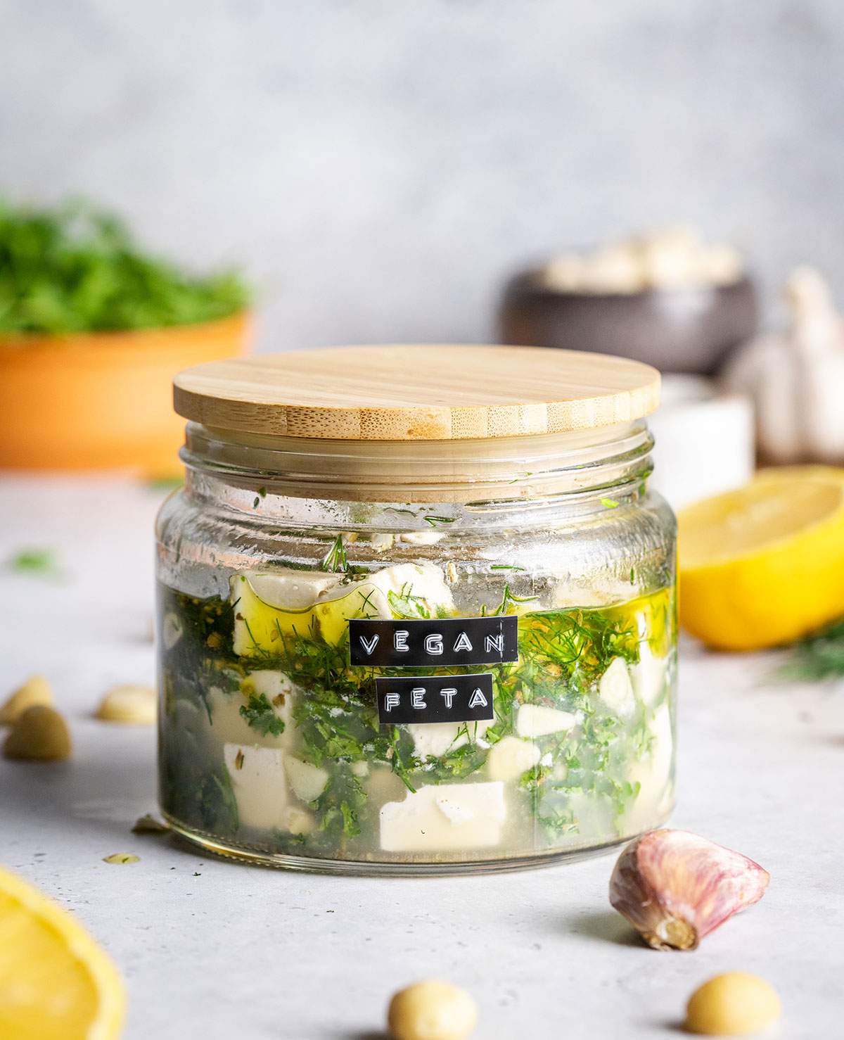 A closed jar filled with vegan feta and marinade is sitting on a white table surrounded by ingredients.