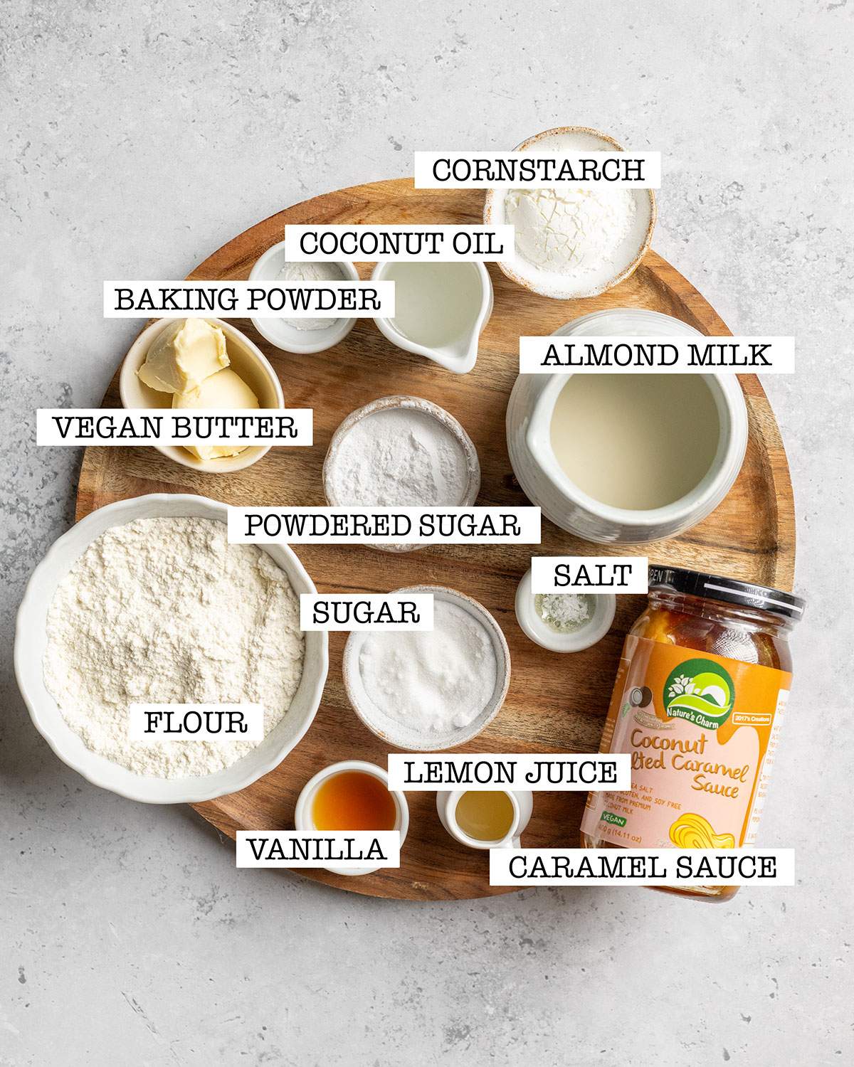 All ingredients for vegan Kaiserschmarrn are shown on a wooden tray with labels naming: flour, sugar, vanilla, lemon juice, caramel sauce, salt, almond milk, powdered sugar, vegan butter, baking powder, coconut oil and cornstarch.