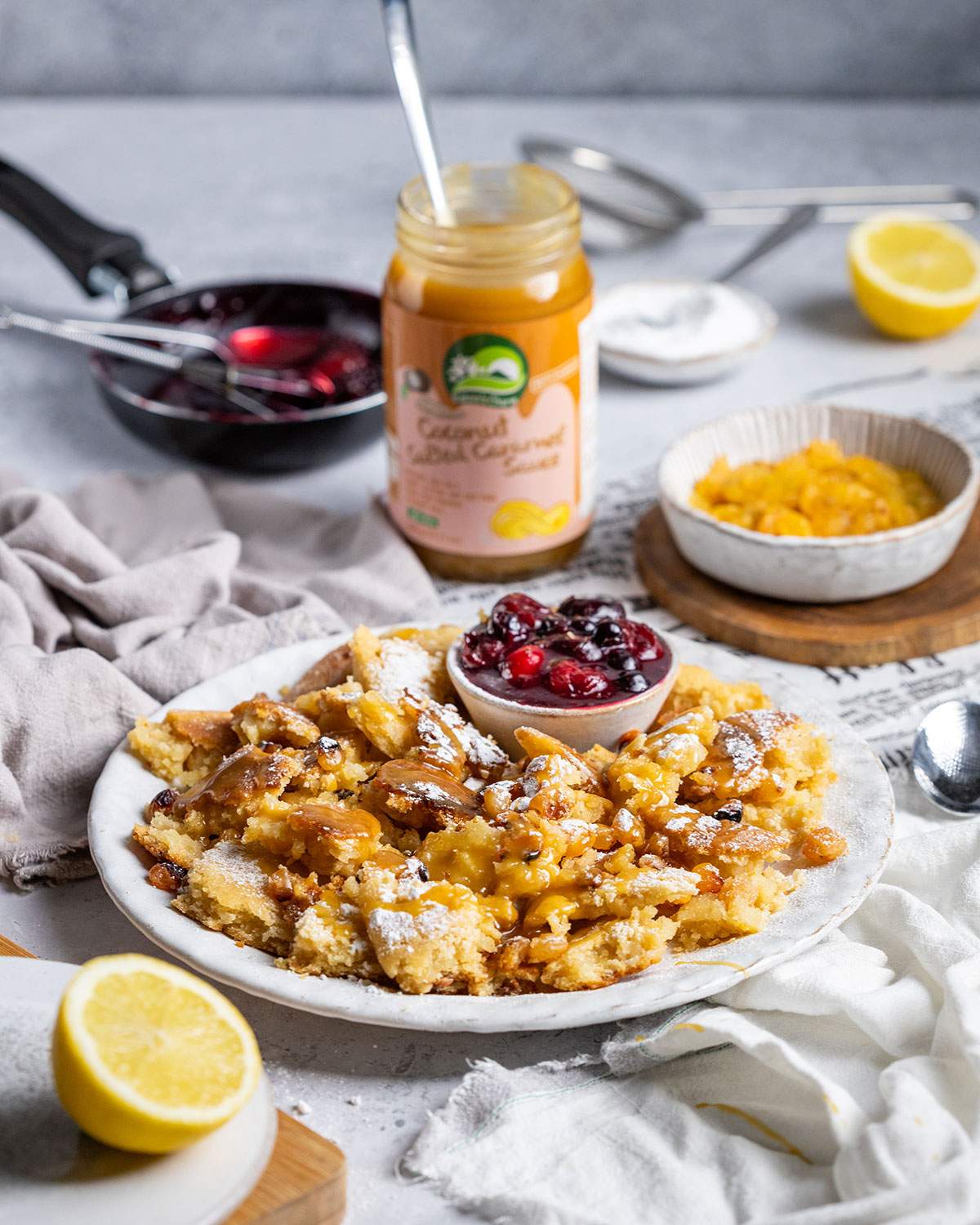 A portion of vegan kaiserschmarrn with cherries is presented on a white plate with caramel sauce drizzled on top.