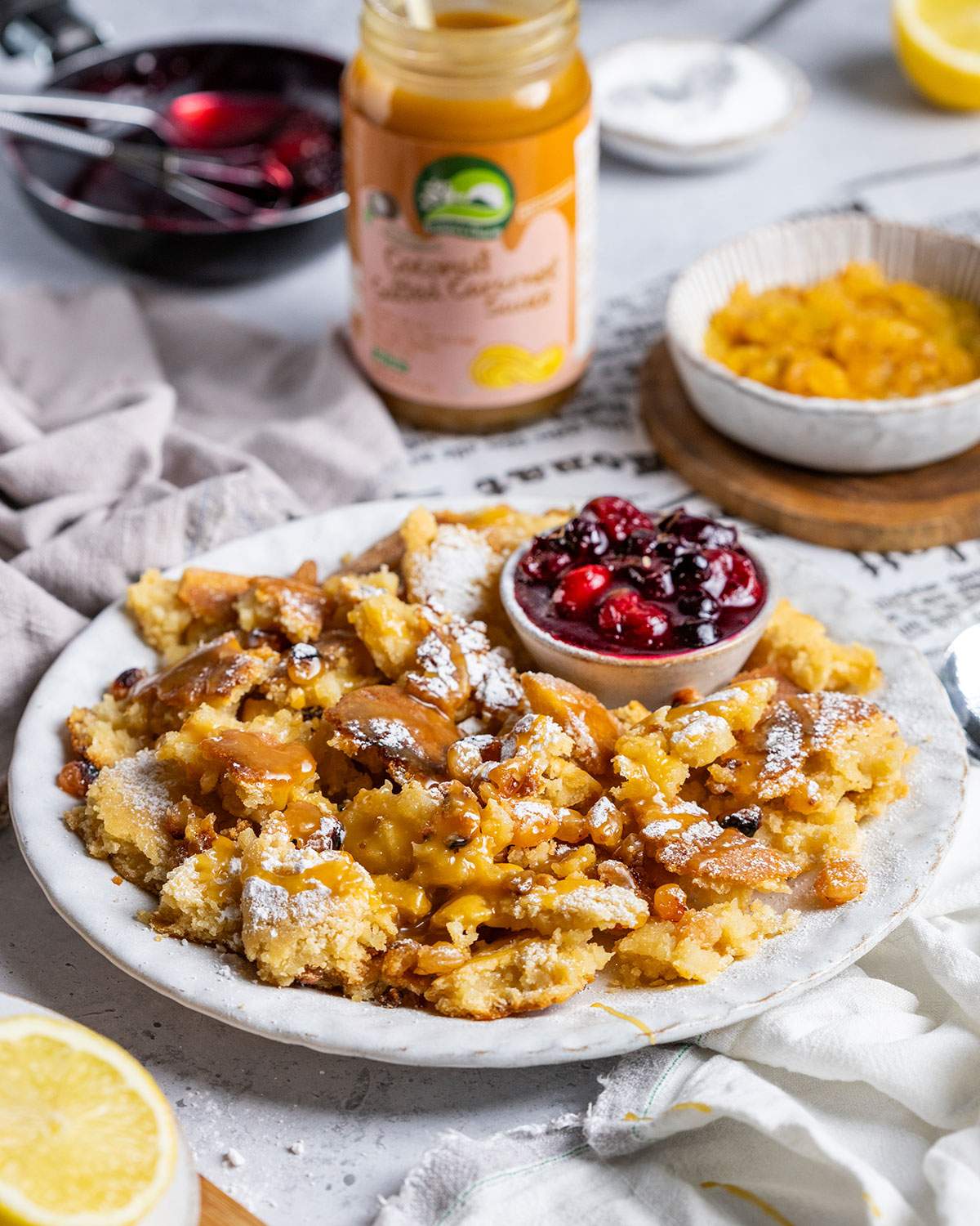 A portion of vegan kaiserschmarrn with cherries is presented on a white plate with caramel sauce drizzled on top.