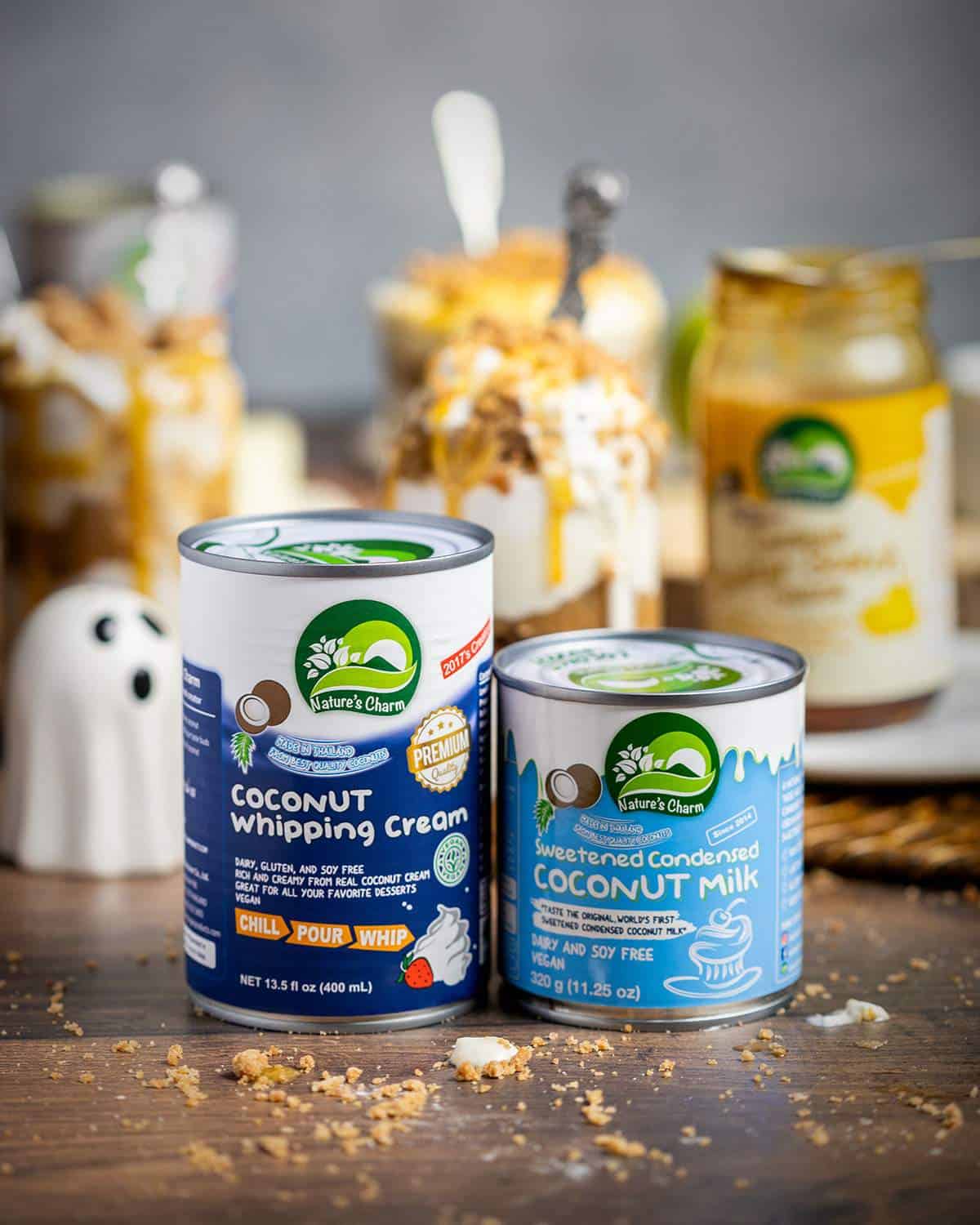 Two cans of Nature's Charm coconut whipping cream and coconut condensed milk on a wooden table with vegan dessert in the background.