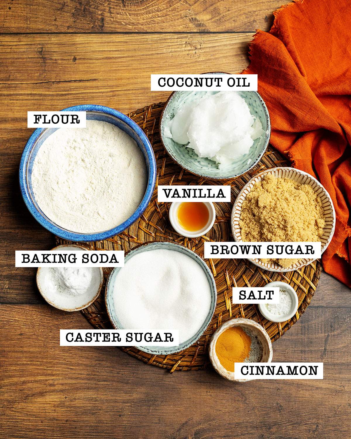 The ingredients for vegan cinnamon cookies are sitting on a round place mat on a wooden table with labels next to each inrgedient.