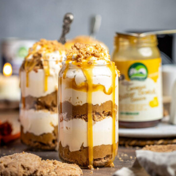 Two dessert glasses filled with vegan caramel apple parfait with layers of caramelized apple, vegan caramel sauce and coconut whipped cream sitting on a wooden table.