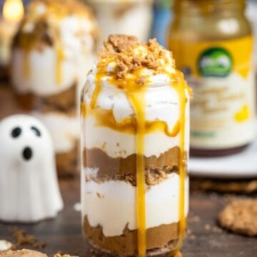 A glass filled to the top with layers layers of caramelised apples, vegan caramel sauce and coconut whipped cream sitting on a wooden table, next to ingredients.