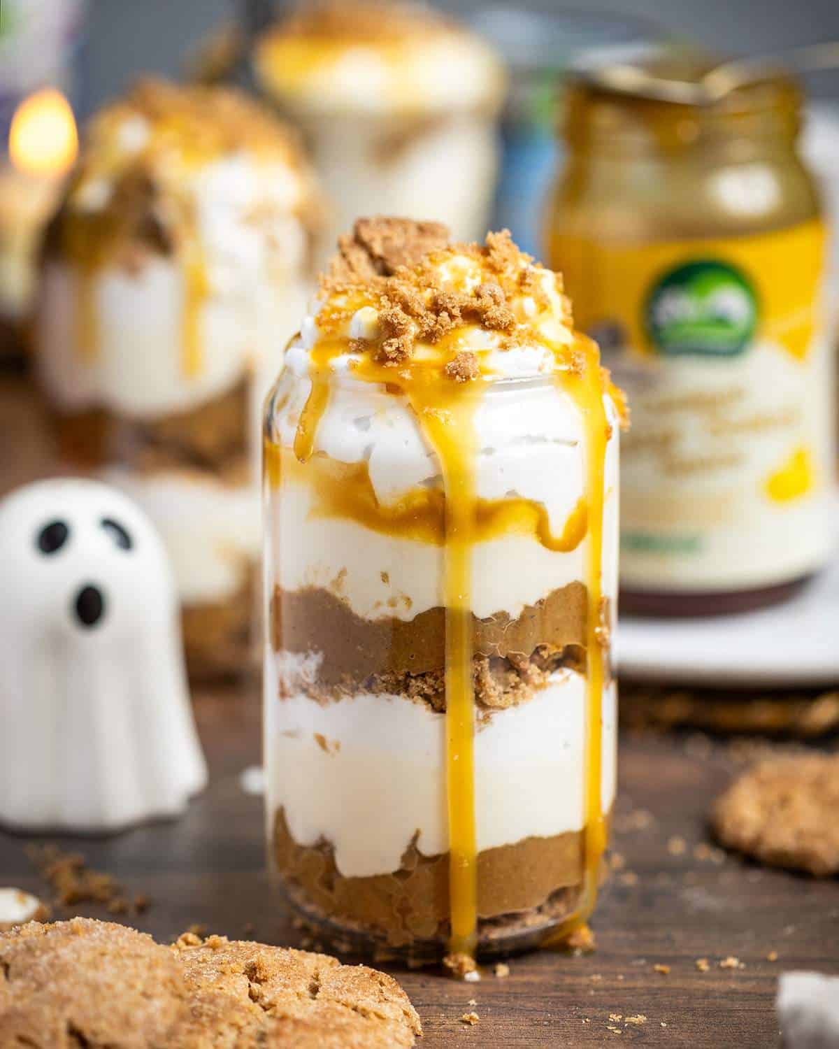 A glass filled to the top with layers layers of caramelised apples, vegan caramel sauce and coconut whipped cream sitting on a wooden table, next to ingredients.