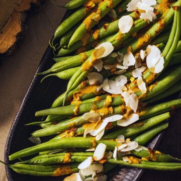 Cooked green beans on a serving platter drizzled with a spicy sauce and almond flakes on top.