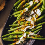Cooked green beans on a serving platter drizzled with a spicy sauce and almond flakes on top.