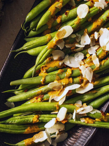 Cooked green beans on a serving platter drizzled with a spicy sauce and almond flakes on top.