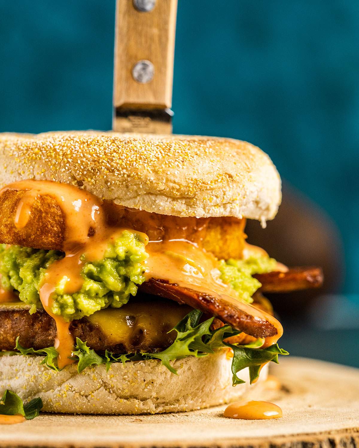 A breakfast burger shown up close with layers of lettuce, English muffin, hashbrown, guacamole, vegan bacon and burger patty, drizzled with a vegan burger sauce.