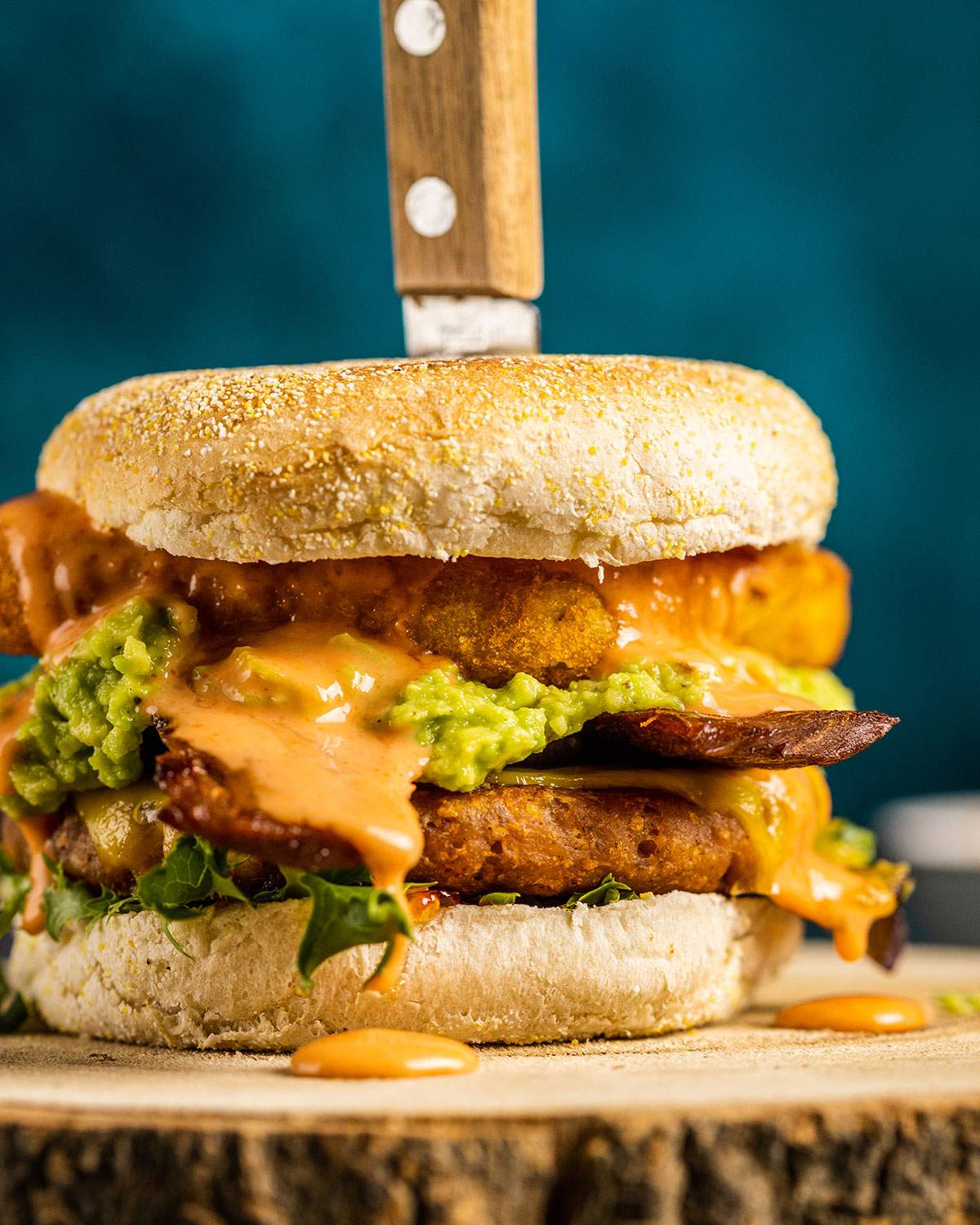 A breakfast burger shown up close with layers of lettuce, English muffin, hashbrown, guacamole, vegan bacon and burger patty, drizzled with a vegan burger sauce.