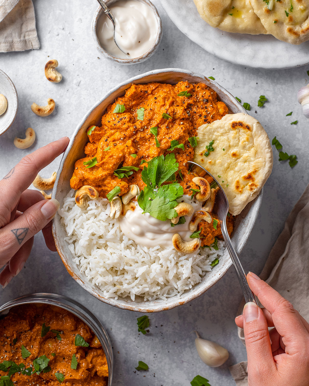 A serving plate with vegan tikka masala alongside basmati rice, fresh cilantro and naan bread on the side. Two hands are holding the plate and one hand is lifting a spoonful of the tikka masala sauce.