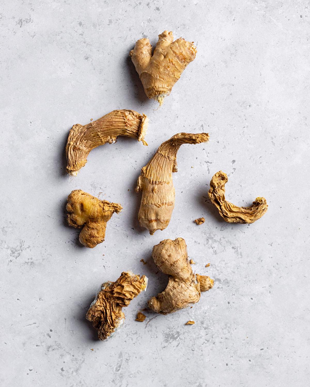 Seven pieces of ginger root at varying stages of quality from fresh to dry, wrinkly and moldy, all sitting on a white table.