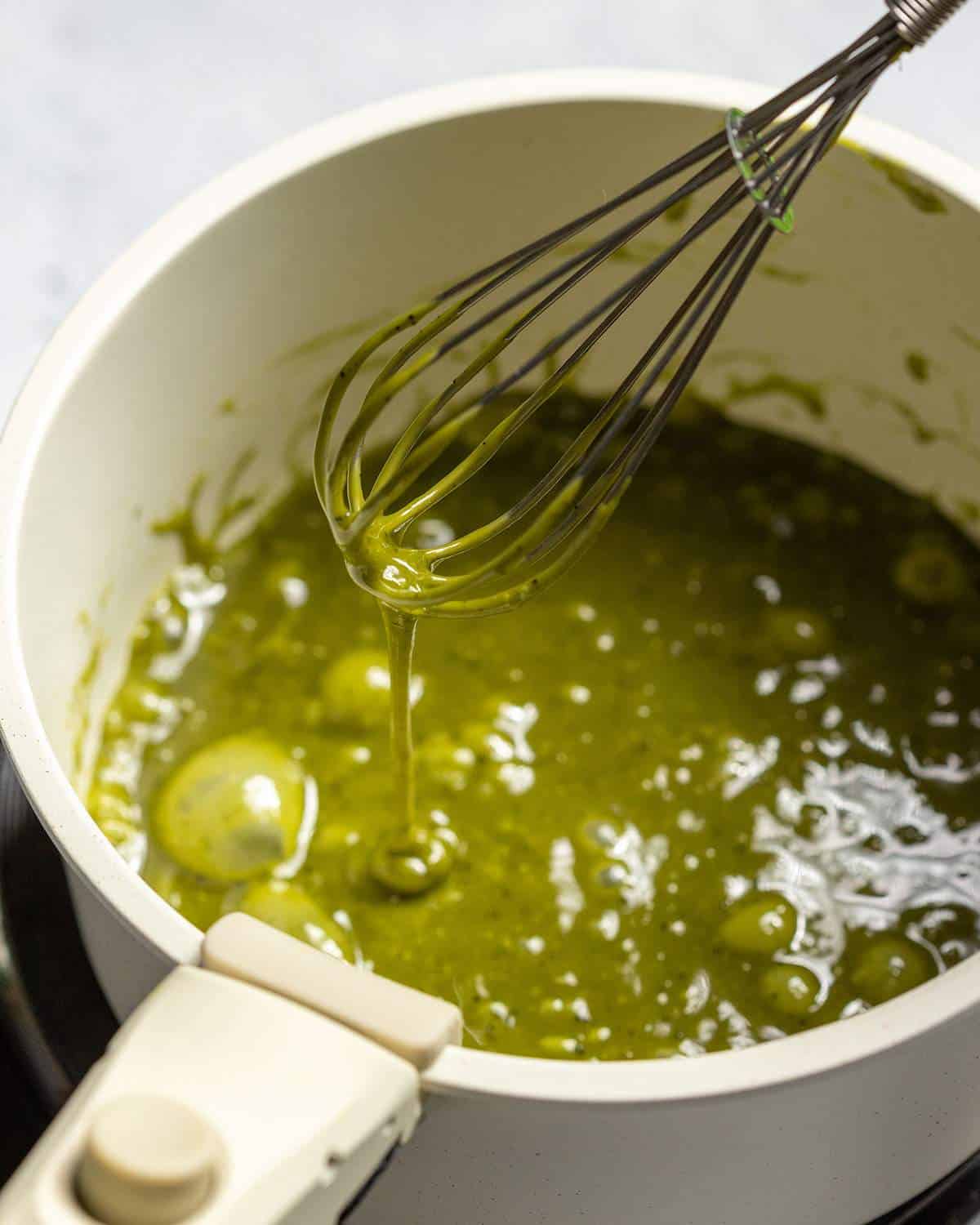 A whisk is lifting matcha caramel out of a saucepan.