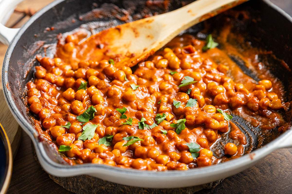 Chickpeas in buffalo sauce are seen in a skillet alongside a wooden spoon.