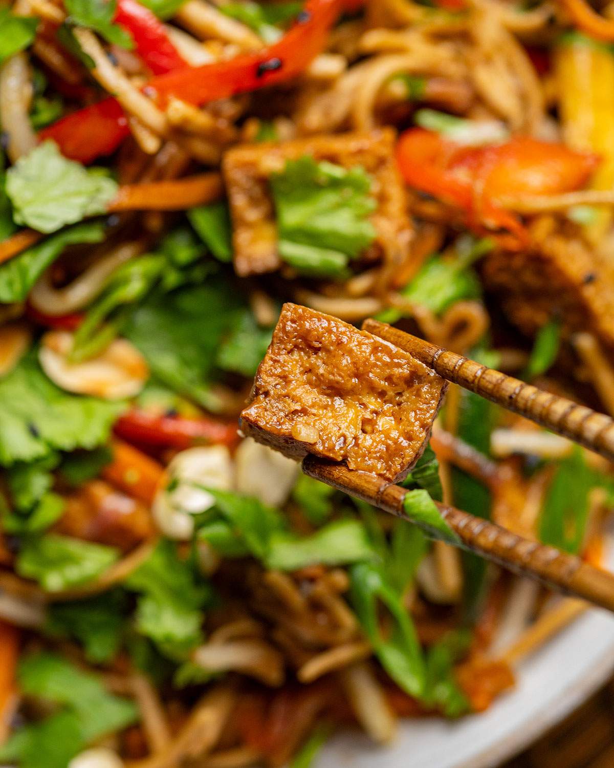 Chopsticks holding a piece of tofu over a bowl of pad thai.