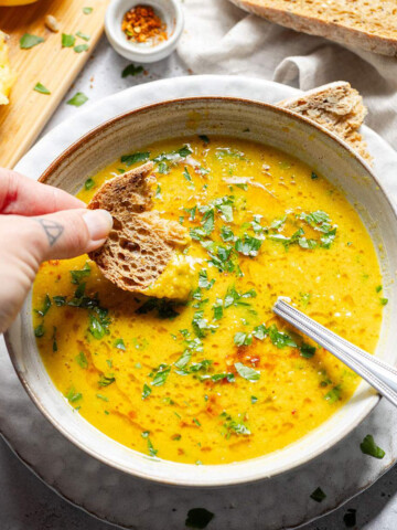 A bowl of hearty lemon soup on a dinner table and a hand dipping a piece of bread into the soup.