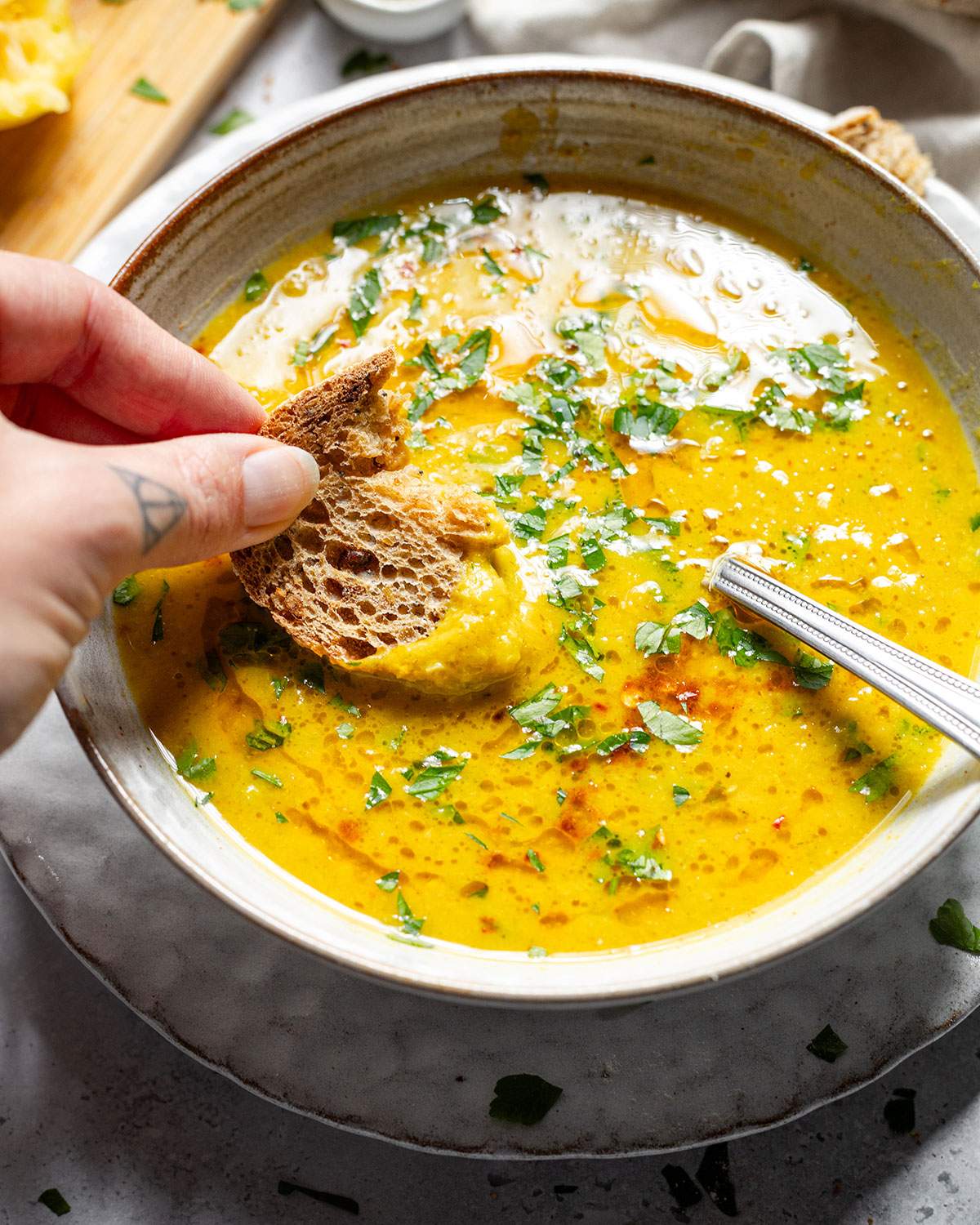 A piece of bread being dipped into a bowl of vibrant lemon soup with fresh herbs on top.