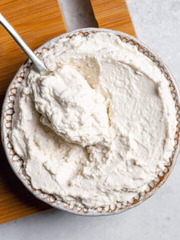 A small bowl of almond ricotta on a serving board with a spoon on the side.