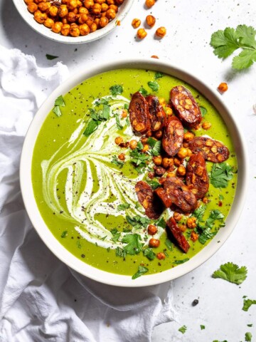 A bowl filled with creamy pea and kale soup, topped with crunchy baked chickpeas and vegan chorizo slices for garnish.