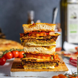 Two focaccia sandwiches layered with crispy aubergine parmigiana, melted vegan cheese, roasted red peppers, and tomatoes on a serving board.