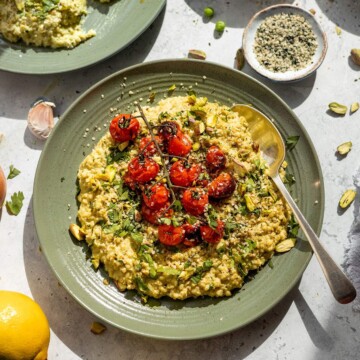 Creamy quinotto aka quinoa risotto served on a green plate with fresh herbs and roasted cherry tomatoes on top. Sat on a table with ingredients surrounding.