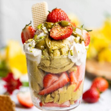 A glass of vegan pistachio ice cream with strawberries, vegan whipped cream and pistachio butter on top.