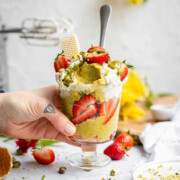 Vegan pistachio ice cream in a jar with strawberries and whipped vegan cream, a hand coming in from the left and picking up the jar.