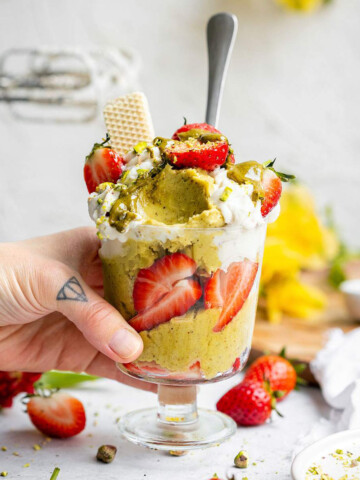 Vegan pistachio ice cream in a jar with strawberries and whipped vegan cream, a hand coming in from the left and picking up the jar.