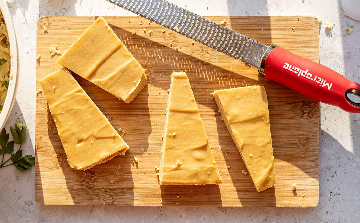 A few wedges of vegan parmesan cheese on a wooden board next to a cheese grater and a pasta dish.