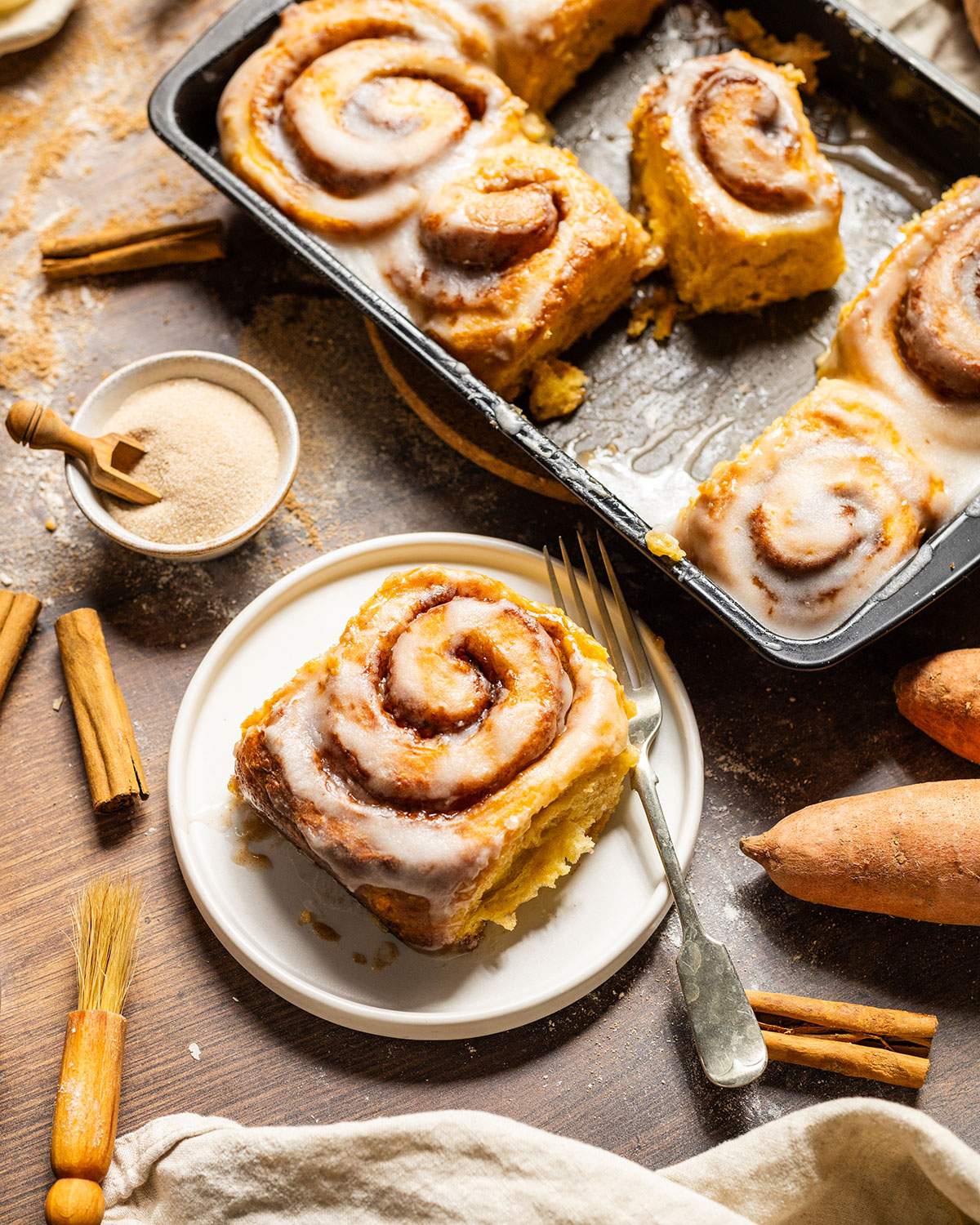 A fluffy sweet potato cinnamon rolls with glaze on a small plate next to the baking dish.