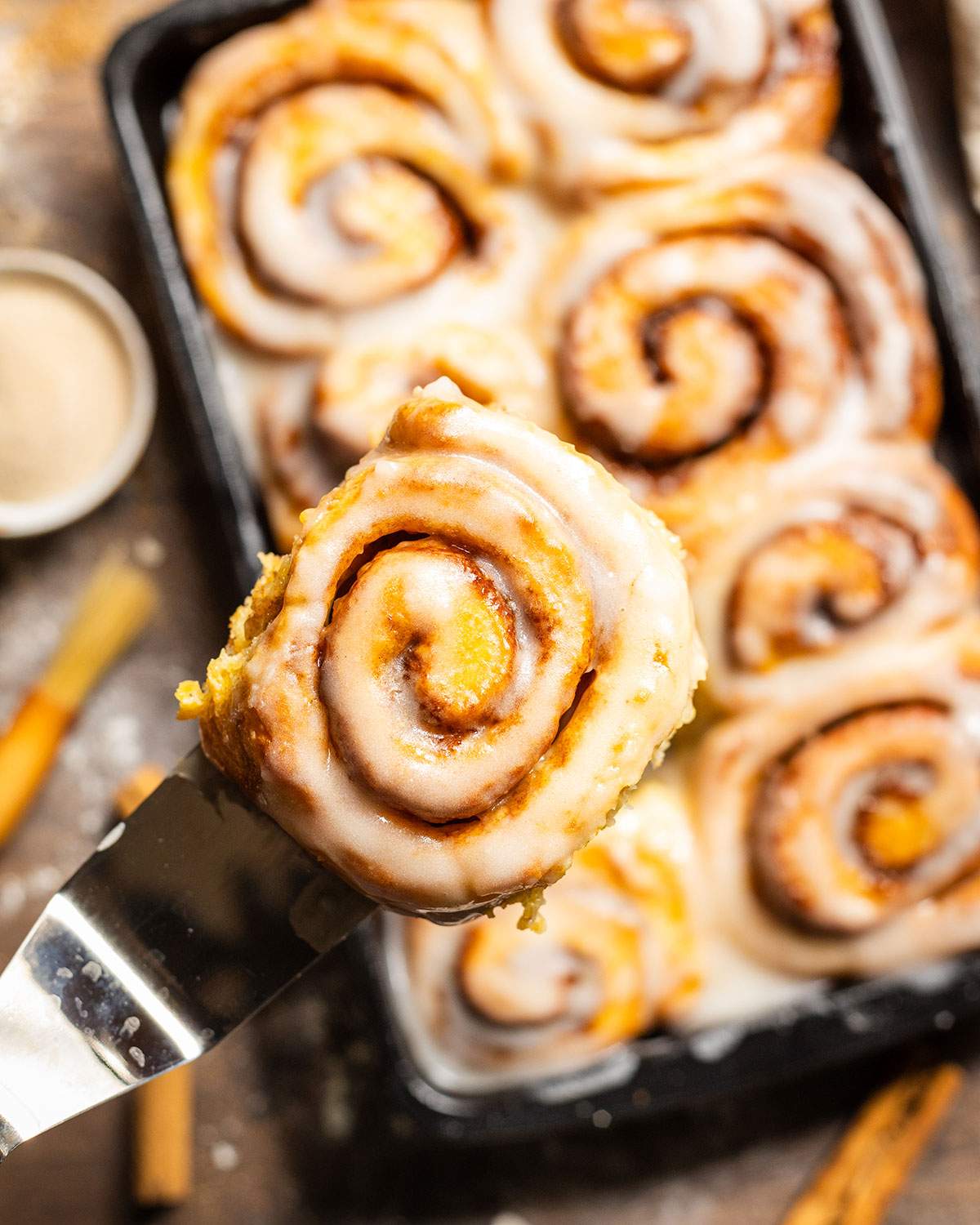A freshly baked sweet potato cinnamon roll held up close above a tray full of rolls.