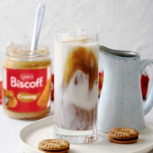 A tall glass with iced biscoff latte on a serving plate with a jar of smooth biscoff spread in the background.