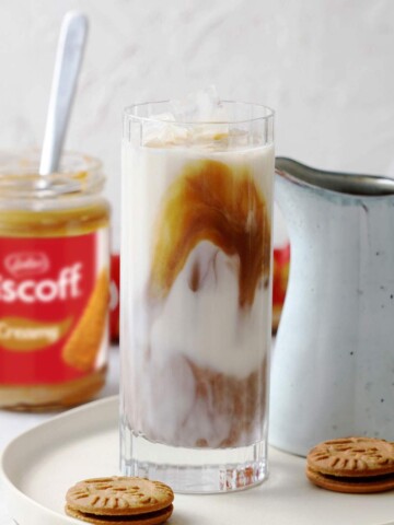 A tall glass with iced biscoff latte on a serving plate with a jar of smooth biscoff spread in the background.