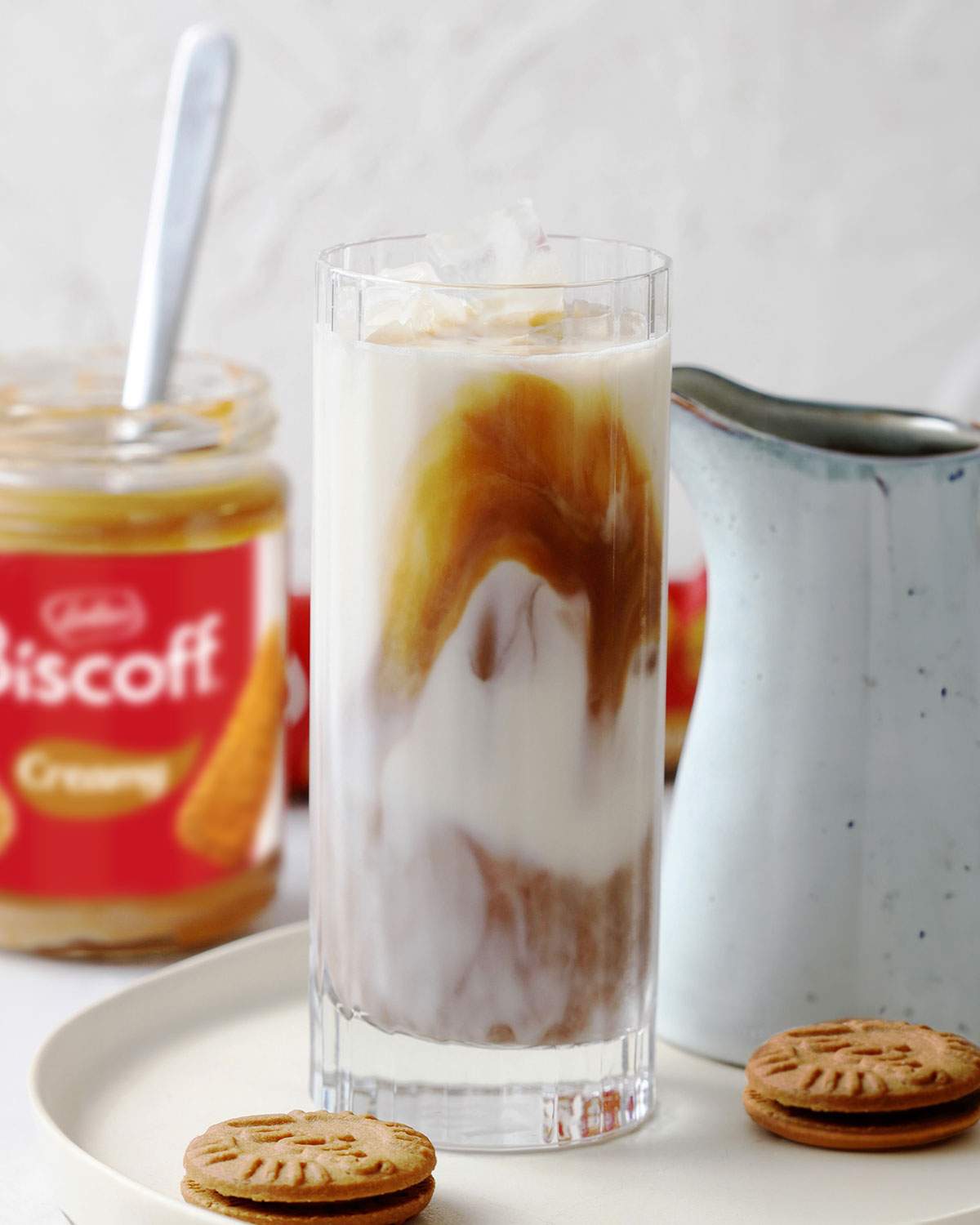 A tall glass with iced biscoff latte on a serving plate with a jar of smooth biscoff spread in the background.